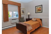 A small, neatly arranged bedroom with a single bed covered in an orange patterned bedspread and a brown throw blanket. There is a wooden nightstand with a lamp and a decorative pillow on the bed. The room has light-colored walls with two framed pictures hanging, a window with orange and brown striped curtains, and a wall-mounted air conditioning unit below the window. The floor is wooden.