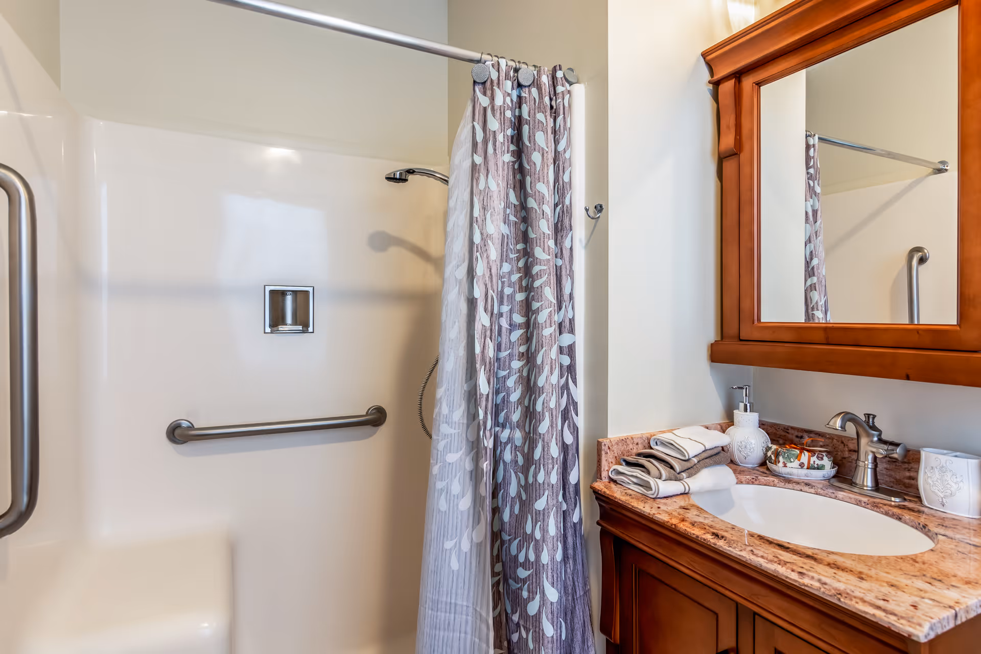 A bathroom with a shower area featuring a grab bar and a shower curtain with a leaf pattern. Next to the shower is a wooden vanity with a granite countertop, a sink, a faucet, a soap dispenser, folded towels, and a large mirror above.