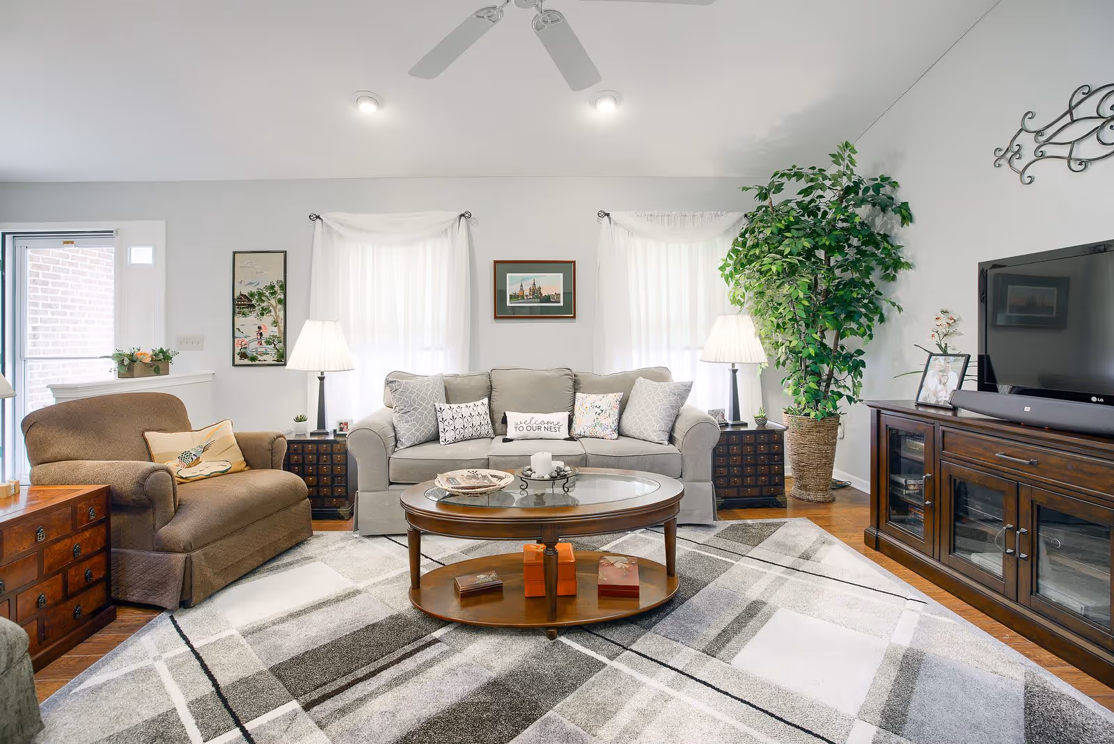 A bright and cozy living room with a beige sofa adorned with multiple pillows, a brown armchair, a round wooden coffee table with decorative items, two side tables with lamps, a large potted plant, a flat-screen TV on a wooden TV stand, and a patterned area rug covering the wooden floor.
