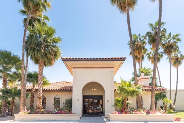 Front entrance of a single-story stucco building with an arched doorway, tiled roof, palm trees, and flowerbeds.