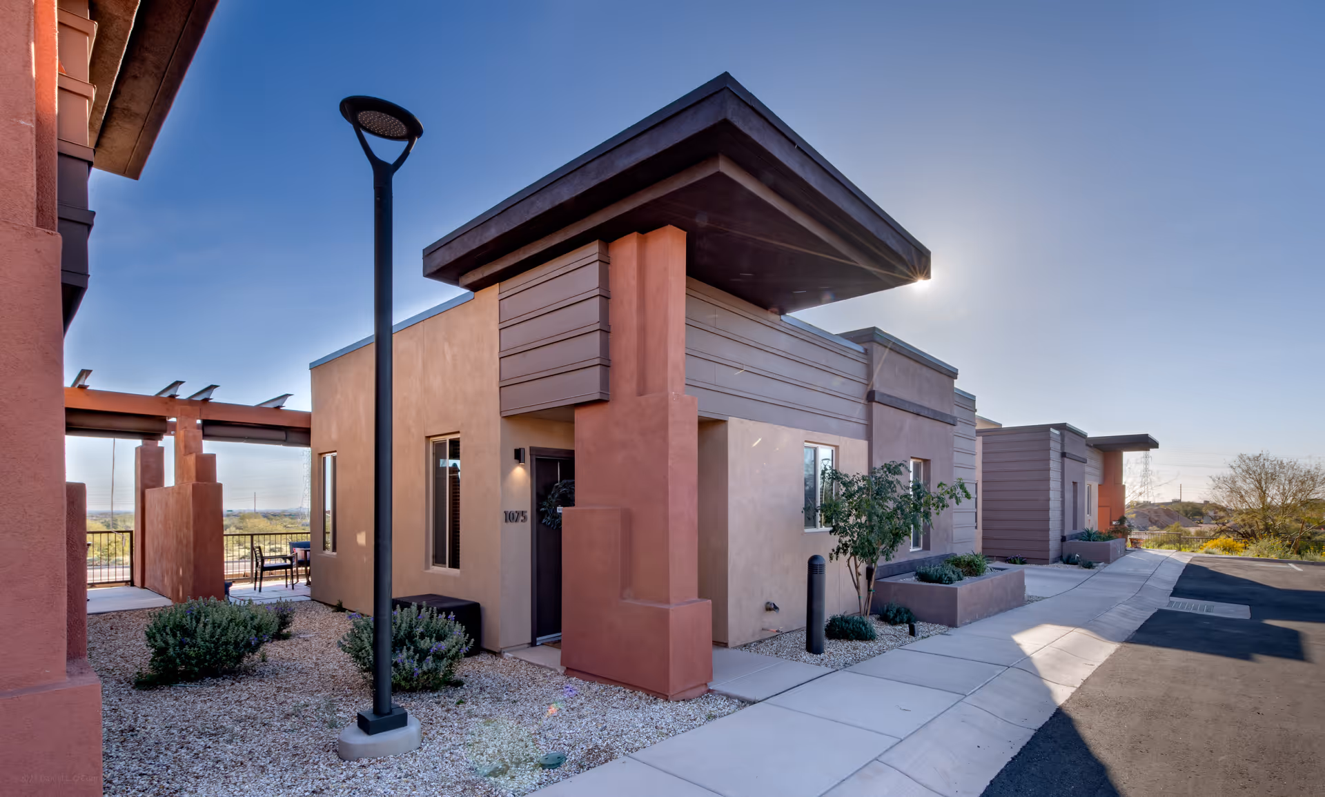 Exterior front view of modern single-story residence units with a covered entry, walkway, desert landscaping, and a streetlamp under a clear blue sky.