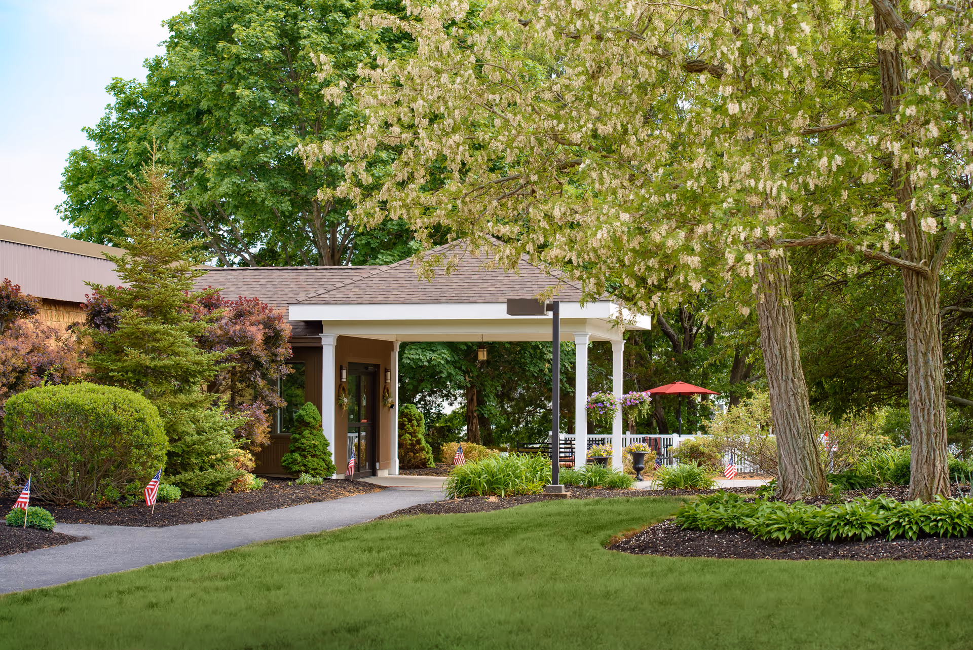 Entrance to a facility surrounded by lush greenery, trees, and well-maintained landscaping with a paved walkway leading to a covered porch area. Small American flags are placed along the garden beds, and a red umbrella is visible in the background.