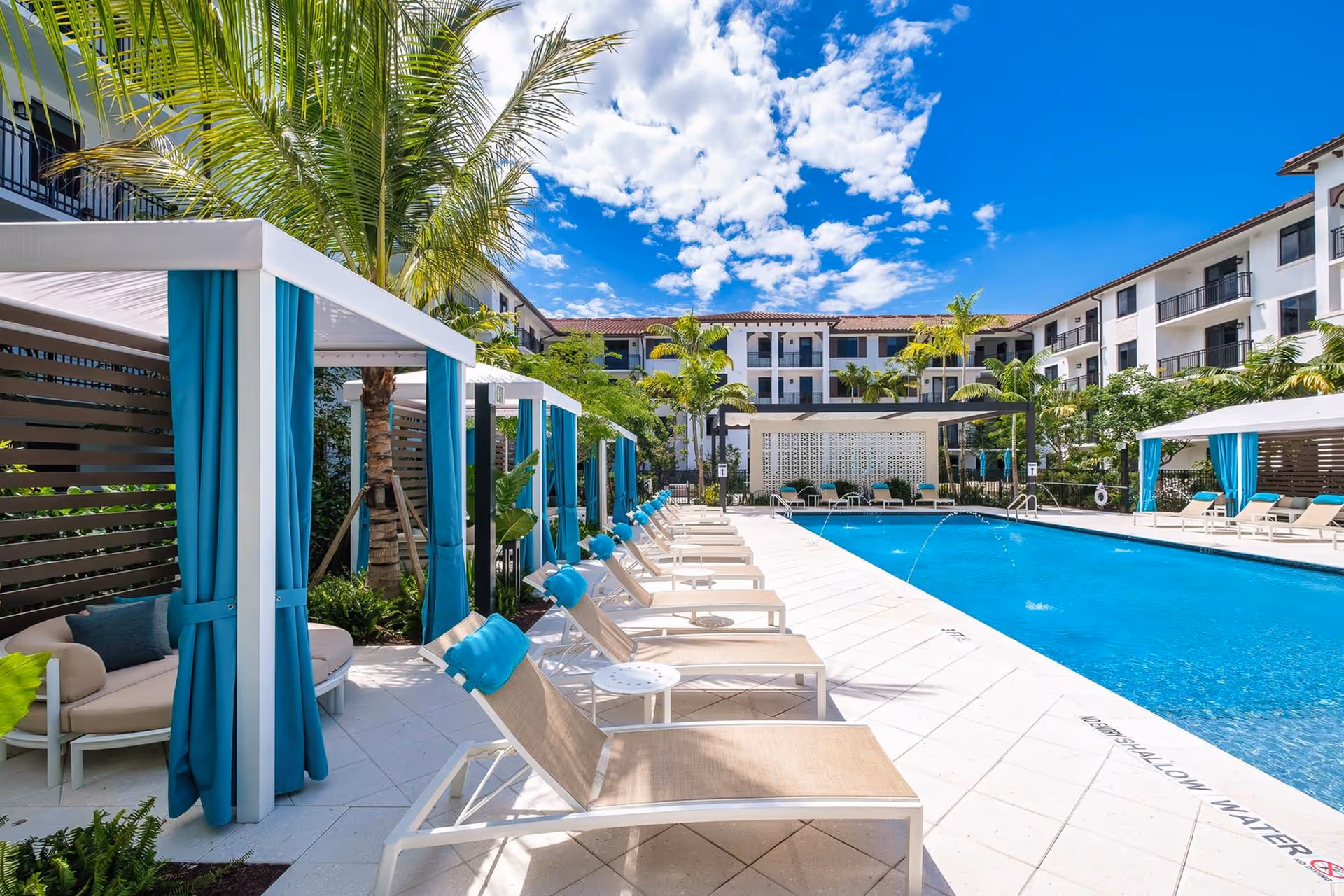 Outdoor swimming pool area at The Residences at Monterra Commons with lounge chairs lined up beside the pool, cabanas with blue curtains, palm trees, and a clear blue sky with scattered clouds. The pool area is surrounded by a multi-story residential building.
