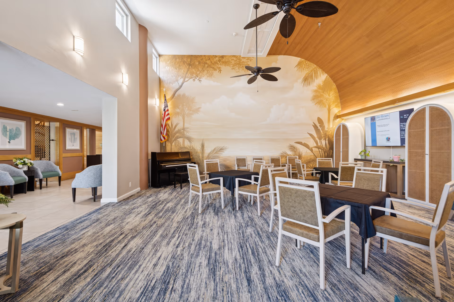 A spacious common area with multiple tables and chairs arranged for group seating. The room features a high ceiling with wooden paneling and two ceiling fans. A large mural depicting a tropical beach scene covers one wall, next to an American flag and a piano. The floor is carpeted with a blue and gray pattern, and there is a large screen mounted on the wall opposite the mural. Adjacent to this room is a seating area with light blue armchairs and framed artwork on the walls.