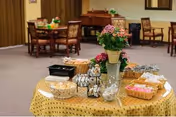 A dining area with a round table covered in a yellow tablecloth displaying various snacks and condiments in baskets and containers. In the background, there are wooden dining tables and chairs, and a piano against the wall.