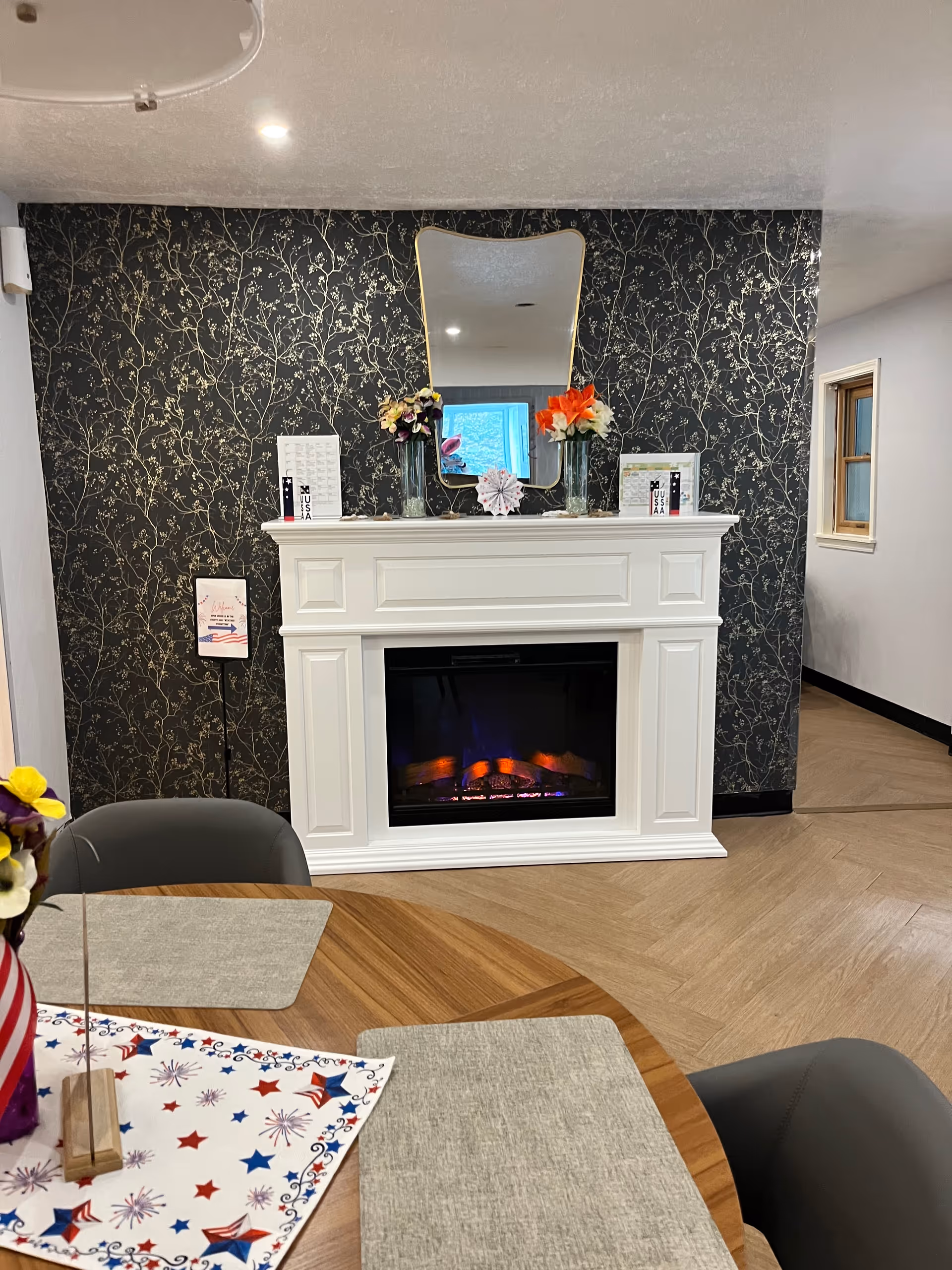A cozy interior scene featuring a white electric fireplace with a decorative mantel against a dark floral wallpapered wall. On the mantel are two vases with colorful flowers, a uniquely shaped mirror, and small framed items. In the foreground is a wooden table with gray placemats and a patriotic-themed table runner with red, white, and blue stars and fireworks. Two gray chairs are partially visible around the table.