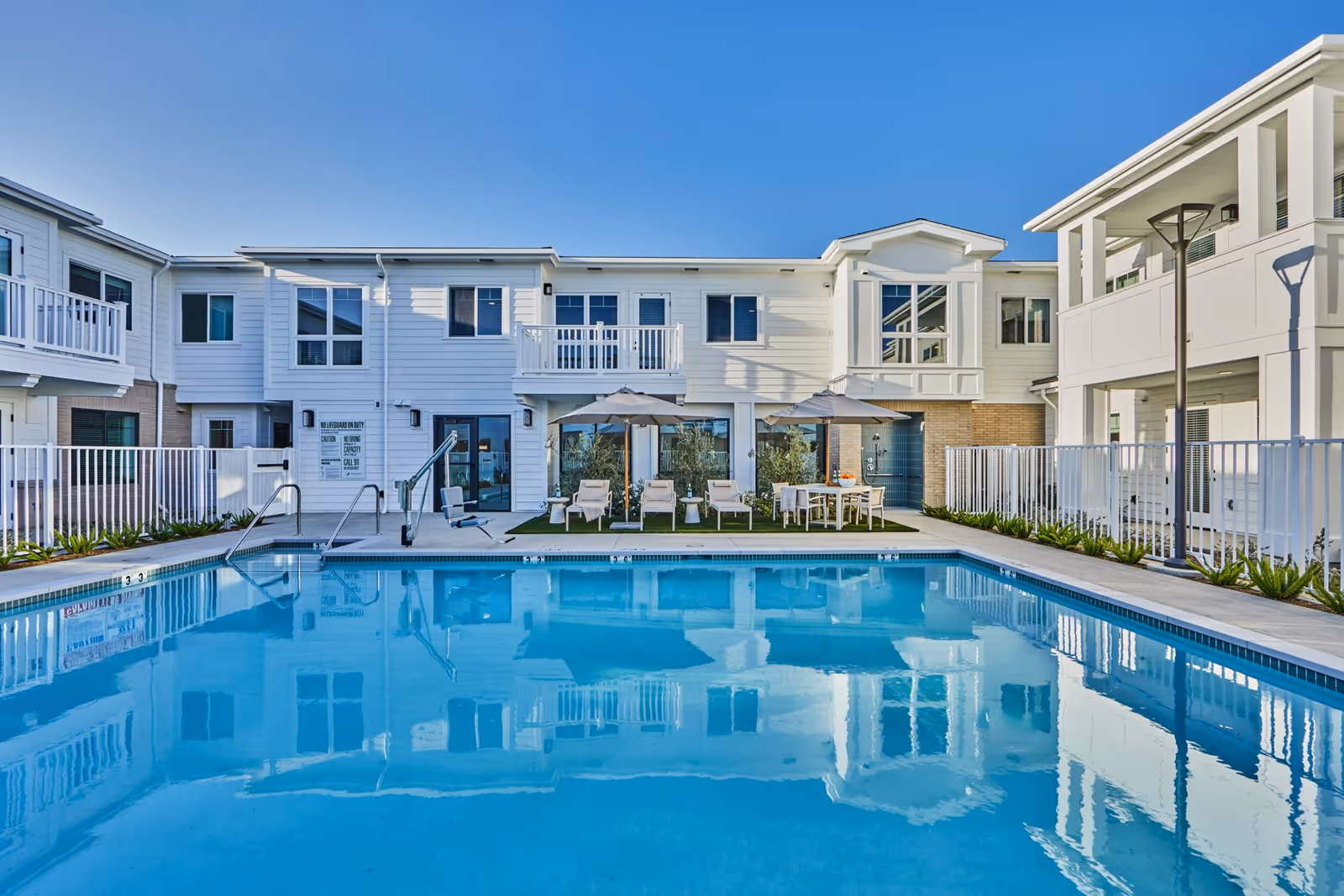 Outdoor swimming pool area at The Seville of San Clemente with clear blue water, surrounded by white fencing and a two-story white building in the background. Poolside features lounge chairs, umbrellas, a table with chairs, and a pool lift for accessibility.