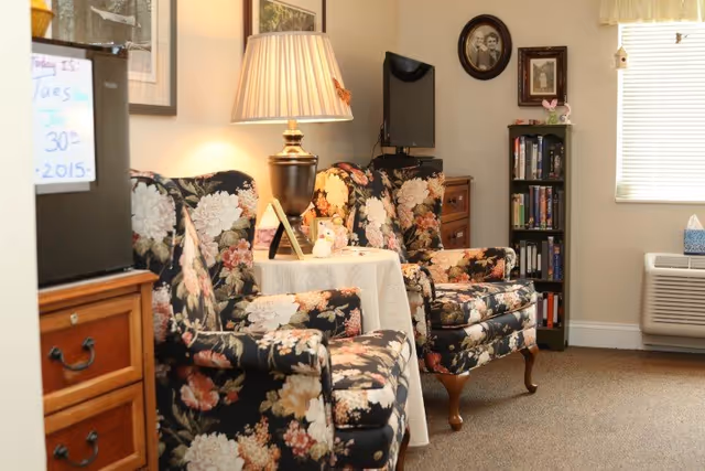 A cozy living room area with two floral upholstered armchairs and a matching sofa arranged around a small round table covered with a white tablecloth. A lamp with a pleated shade sits on the table. The room has framed pictures on the walls, a small bookshelf filled with books and DVDs, a flat-screen TV mounted on the wall, and a window with blinds partially open. There is also a wooden dresser and an air conditioning unit below the window.