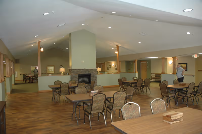 Spacious common room with multiple tables and chairs, a central stone fireplace, and wood flooring.