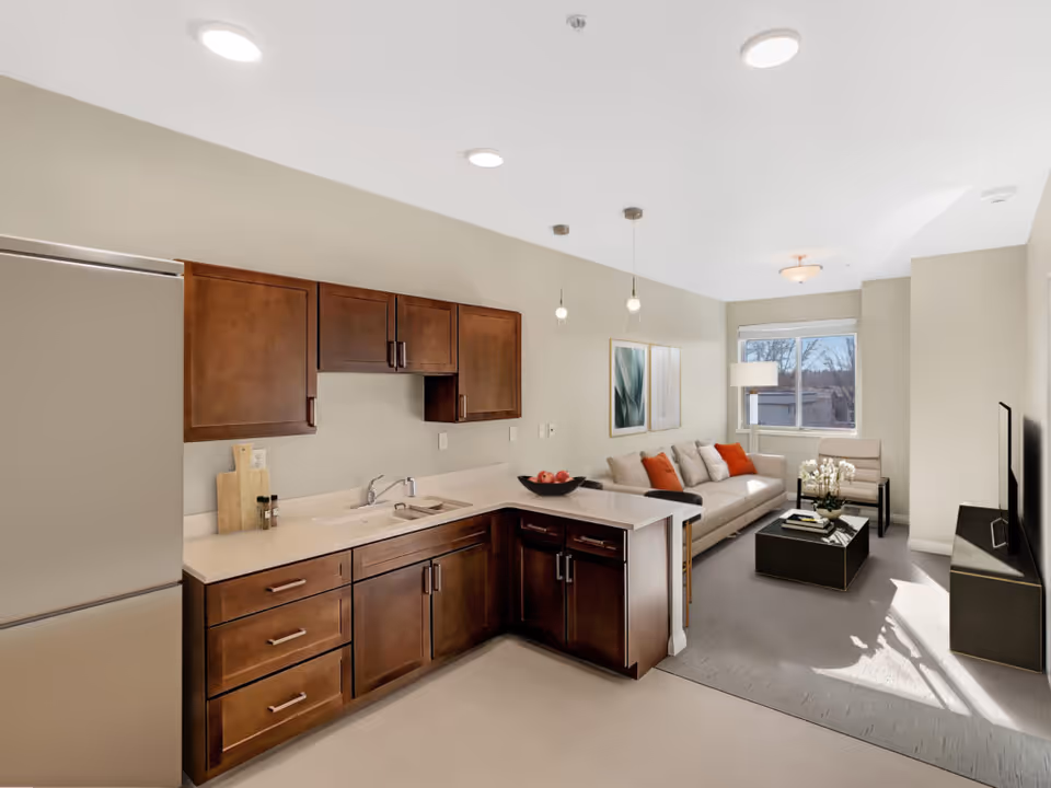Open-plan kitchen and living area with dark wood cabinets, a beige sofa with orange pillows, and a TV near a window.