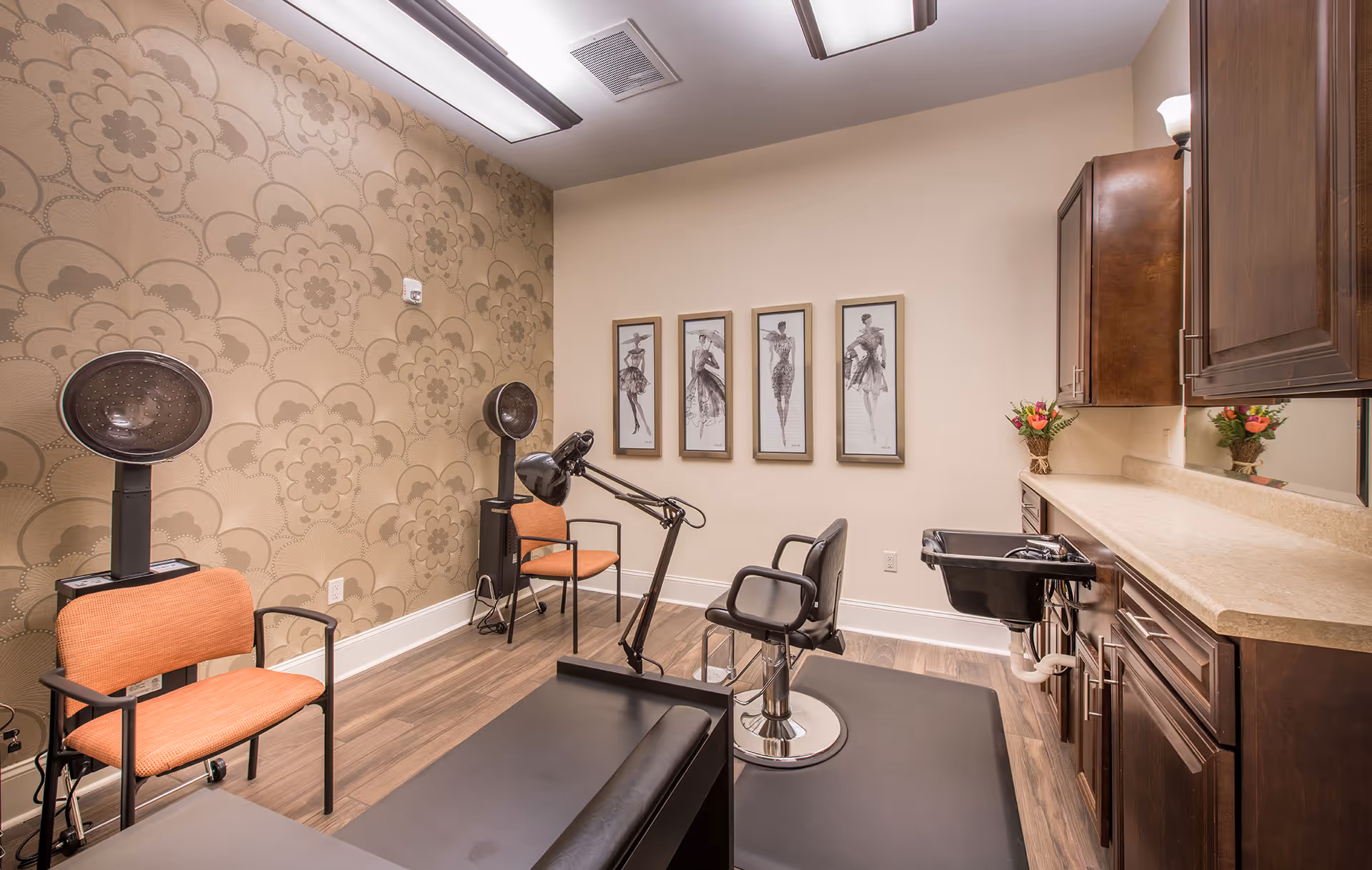 Interior view of a salon room with a hair washing station, a black salon chair on a black mat, two orange chairs with hair dryers behind them, a countertop with dark wooden cabinets above and below, floral wallpaper on one wall, and four framed fashion sketches on the opposite wall.