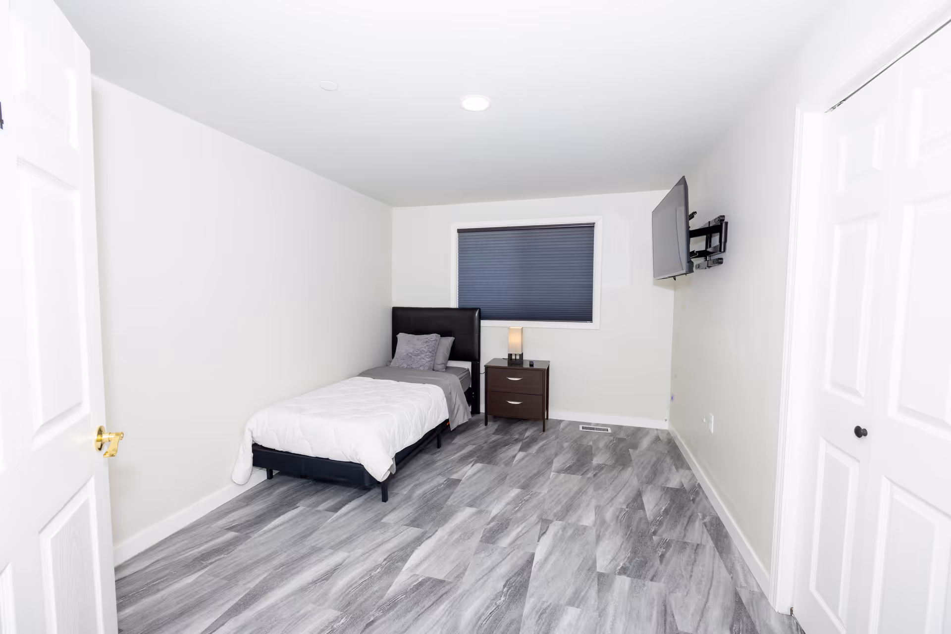 A simple, modern bedroom with a single bed against the left wall, a nightstand with a lamp next to it, a window with a dark blind, and a wall-mounted TV on the right. The room has light-colored walls and gray wood-patterned flooring.