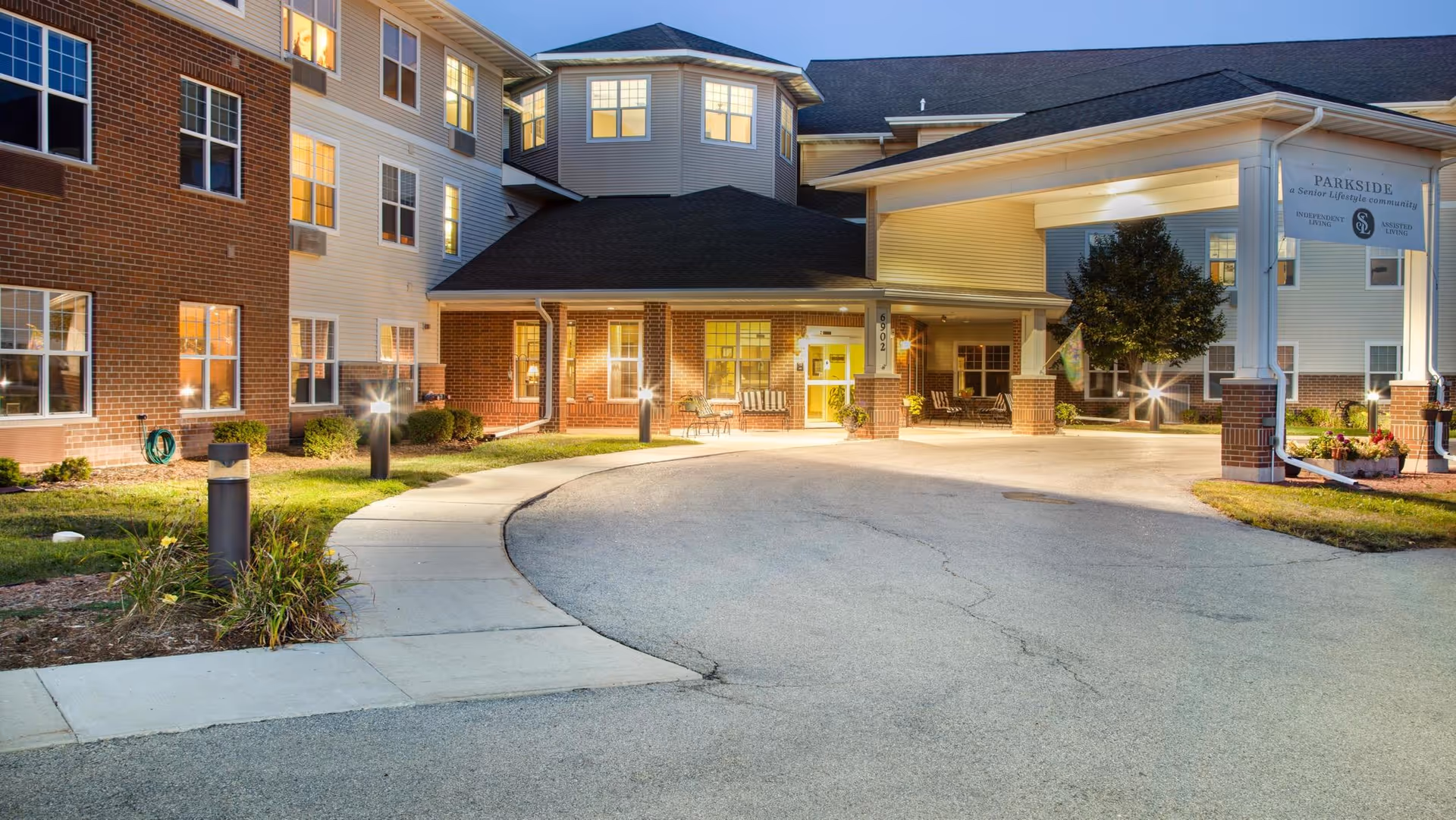 Exterior view of Parkside Senior Living facility at dusk, showing a well-lit entrance with a covered driveway, brick and siding facade, multiple windows with lights on inside, landscaped greenery, and a sign displaying the facility's name.