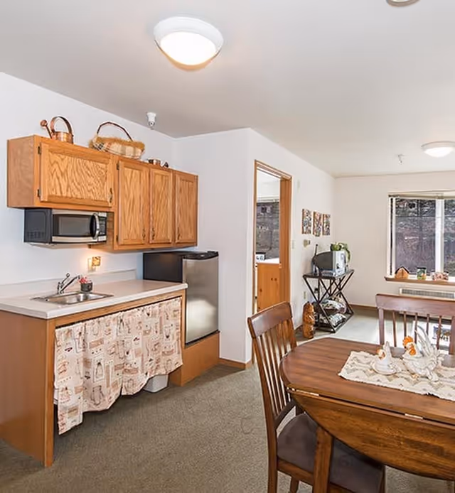 A small kitchen area with wooden cabinets, a microwave, a sink, and a mini refrigerator. There is a dining table with chairs in the foreground, and a window with a view outside in the background. The room has light-colored walls and carpeted flooring.