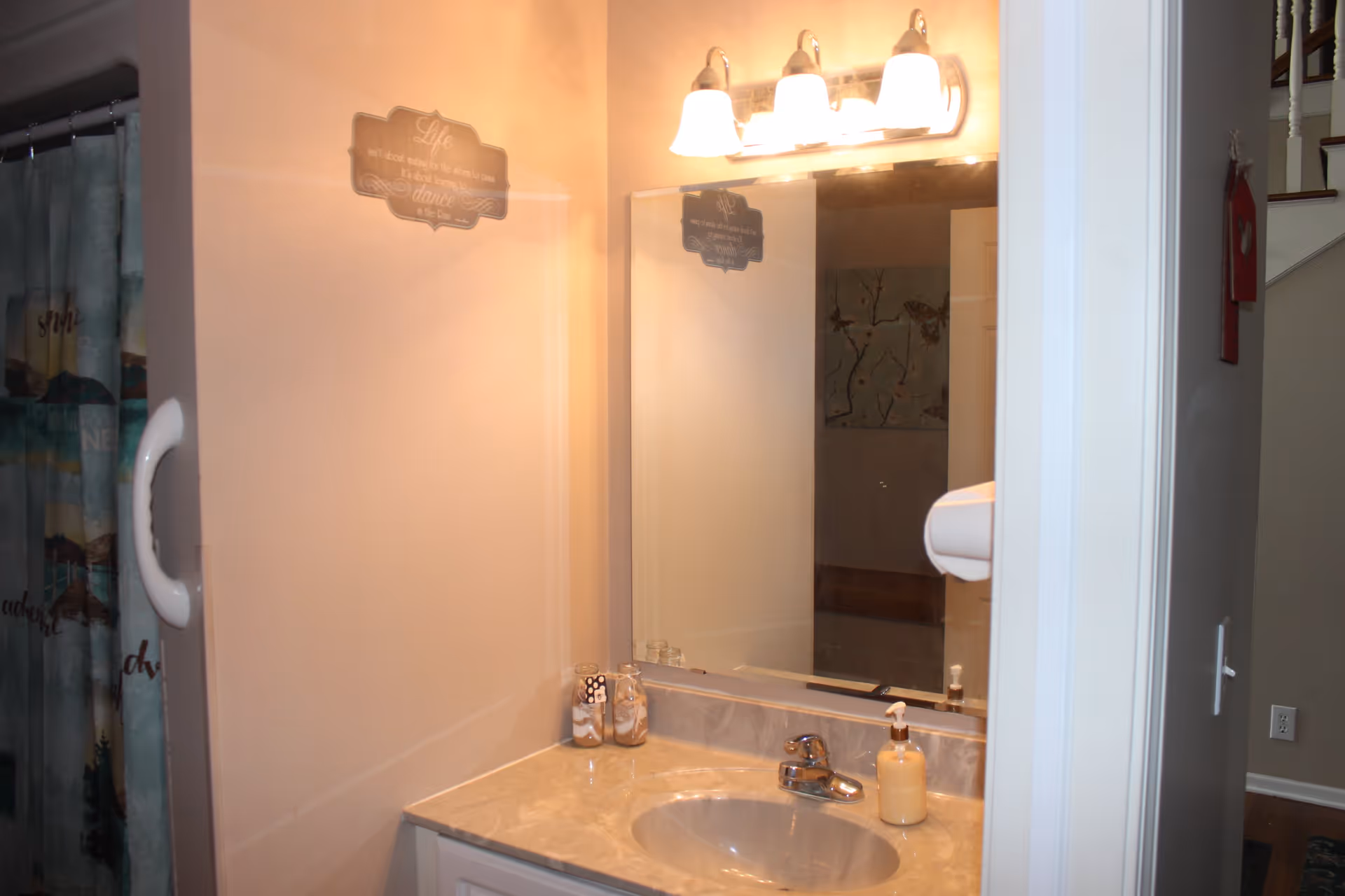 Bathroom sink with a marble countertop, a faucet, a soap dispenser, and two decorative jars. Above the sink is a large mirror with a three-light fixture. To the left, a shower curtain with a nature-themed design is partially visible. The wall has a decorative sign with an inspirational quote.