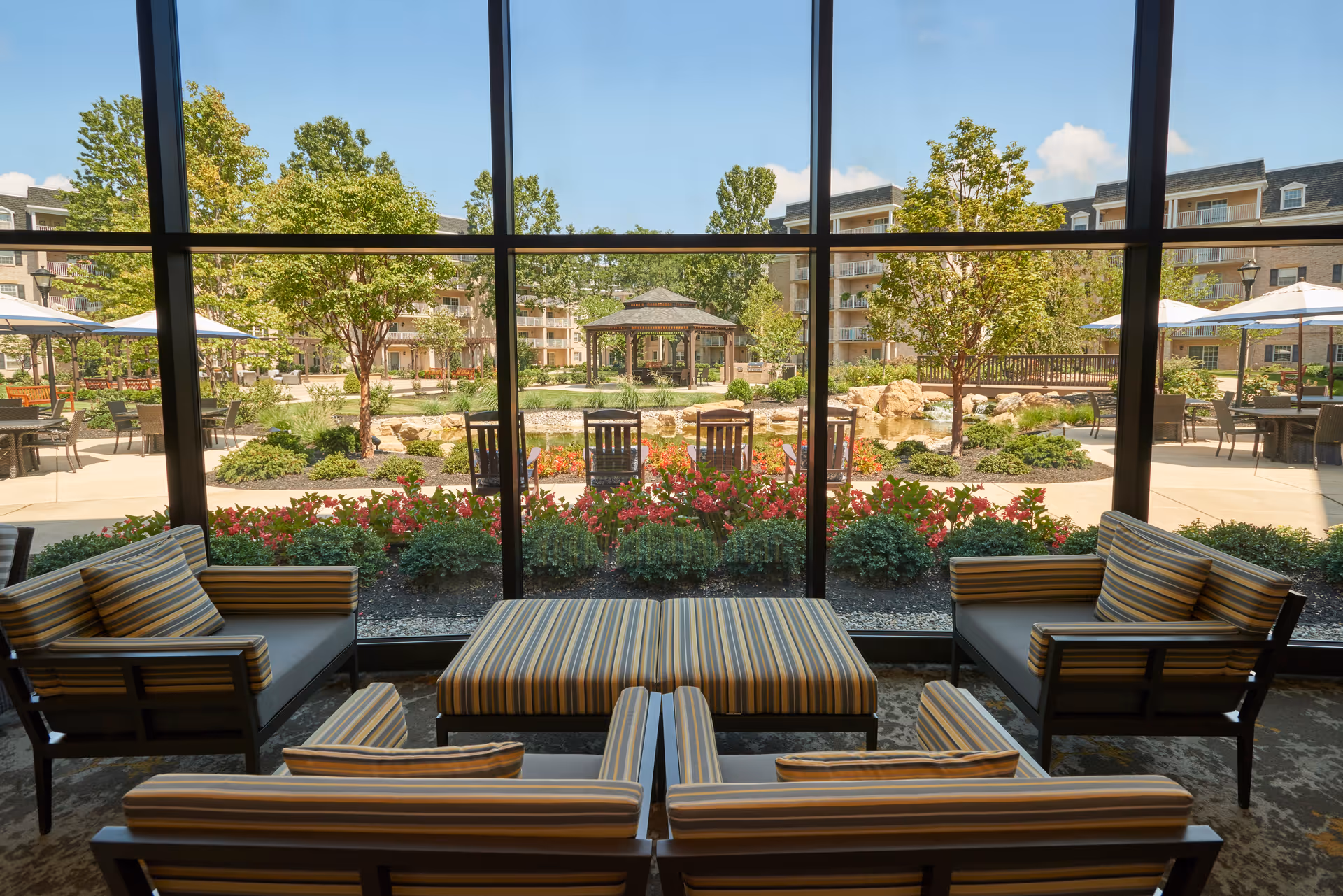 View from inside a lounge area with striped cushioned chairs and an ottoman, looking out through large floor-to-ceiling windows onto a landscaped outdoor garden with trees, flowers, a gazebo, and patio seating with umbrellas.