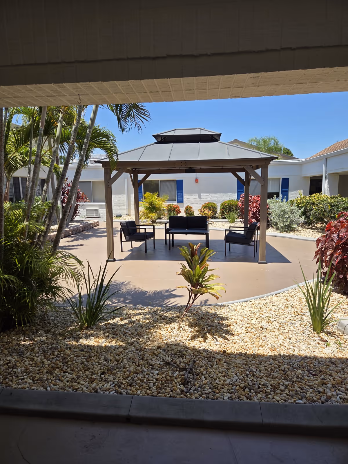 Outdoor courtyard area with a covered gazebo featuring a seating arrangement of a couch and three chairs. The area is surrounded by small plants, bushes, and palm trees, with a clear blue sky overhead. The courtyard is part of a building with white walls and blue window shutters.