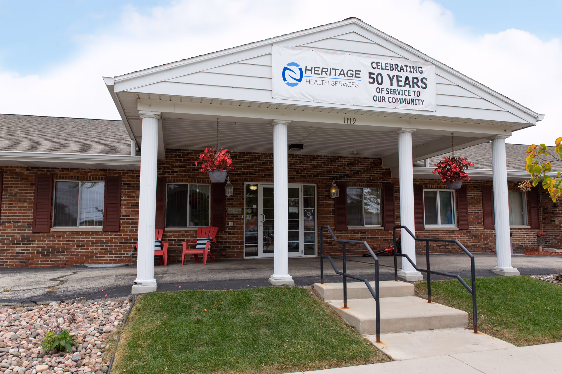 Front entrance of the Heritage Health Services brick building with white columns, a banner celebrating 50 years, hanging flower baskets, and chairs on the porch.