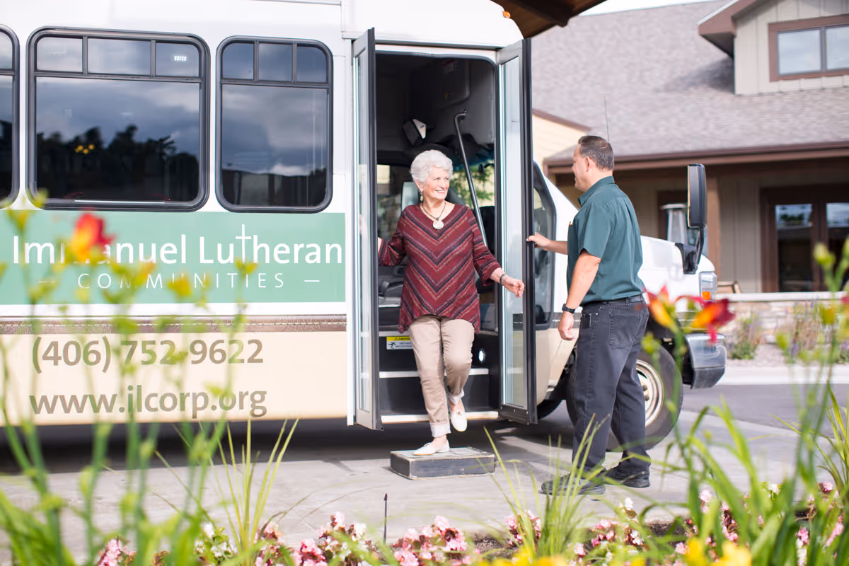 An elderly woman steps down from a shuttle bus labeled Immanuel Lutheran Communities, assisted by a man standing outside. The scene is outdoors with flowers in the foreground and a building in the background.