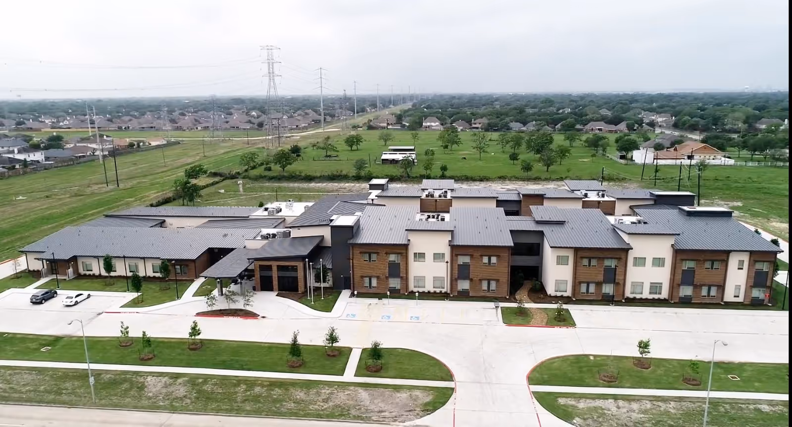 Aerial view of a large, modern senior living facility building with multiple sections and a gray roof, surrounded by green lawns and a few parked cars. The facility is located in a suburban area with houses and open fields in the background under a cloudy sky.