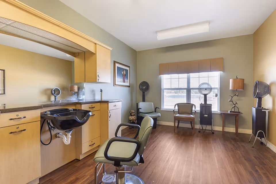 A bright hair salon room with wooden flooring and light green walls. The room features a black salon sink with a mirror and wooden cabinets, two green salon chairs, two vintage hair dryers, a wooden chair, a small wooden table with a lamp, and a large window with blinds letting in natural light.