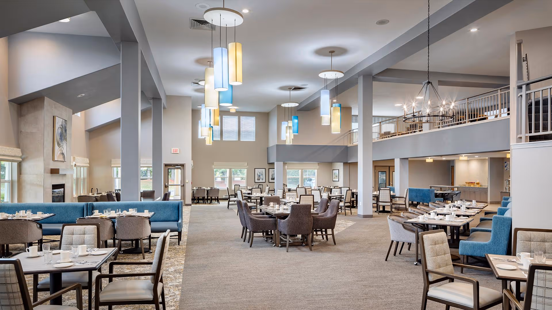 Spacious and well-lit dining area in Holiday White Oaks facility featuring multiple tables and chairs arranged neatly, modern pendant lighting in blue, yellow, and white hues hanging from the ceiling, large windows allowing natural light, and a cozy fireplace on the left side.