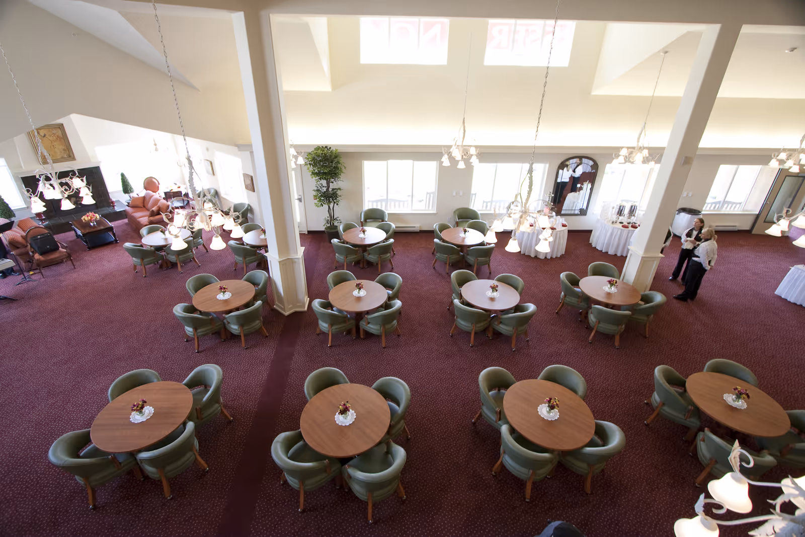 A spacious dining room with multiple round wooden tables each surrounded by four green cushioned chairs. The room has a maroon carpet and is well-lit with hanging chandeliers. There are large windows along the far wall letting in natural light. In the background, there is a seating area with sofas and two people standing near a table with white tablecloths and refreshments.