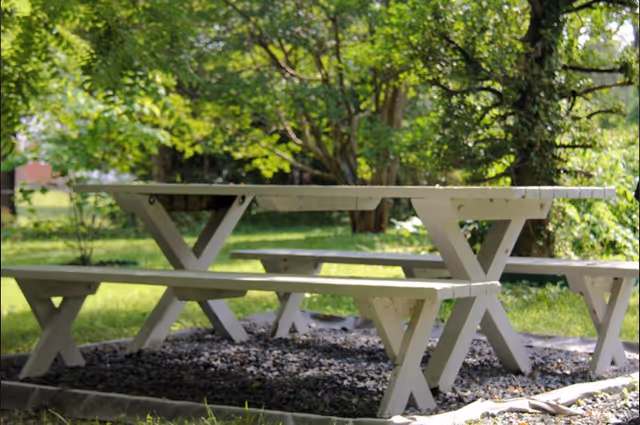 A white wooden picnic table with attached benches situated outdoors on a bed of small dark stones, surrounded by green grass and trees in the background.