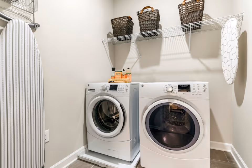 A front-loading washer and dryer in a small laundry room with an ironing board, wire shelf with baskets, and detergent on top.