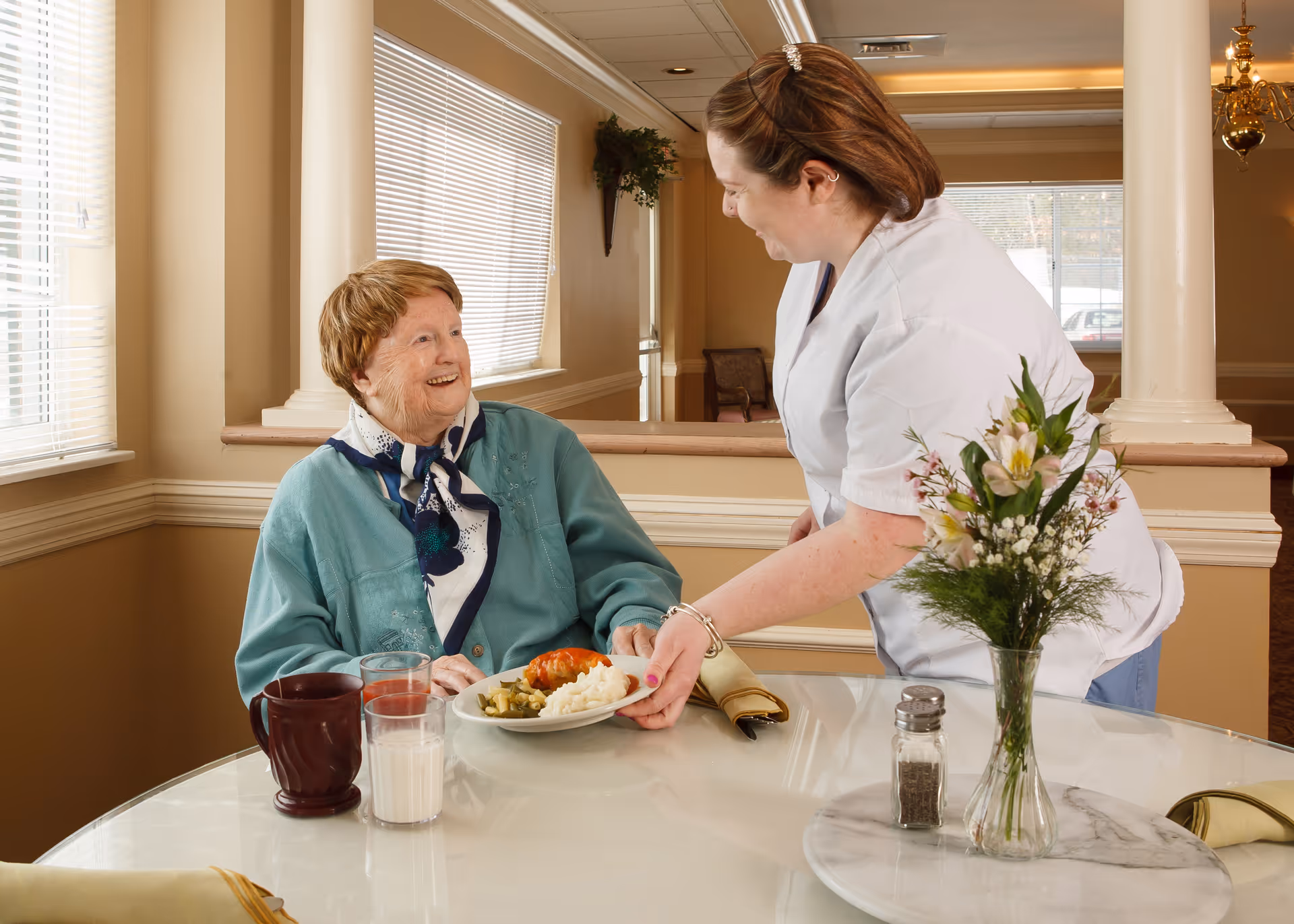 A caregiver serves a smiling elderly woman a plate of food at a dining table decorated with a vase of flowers.