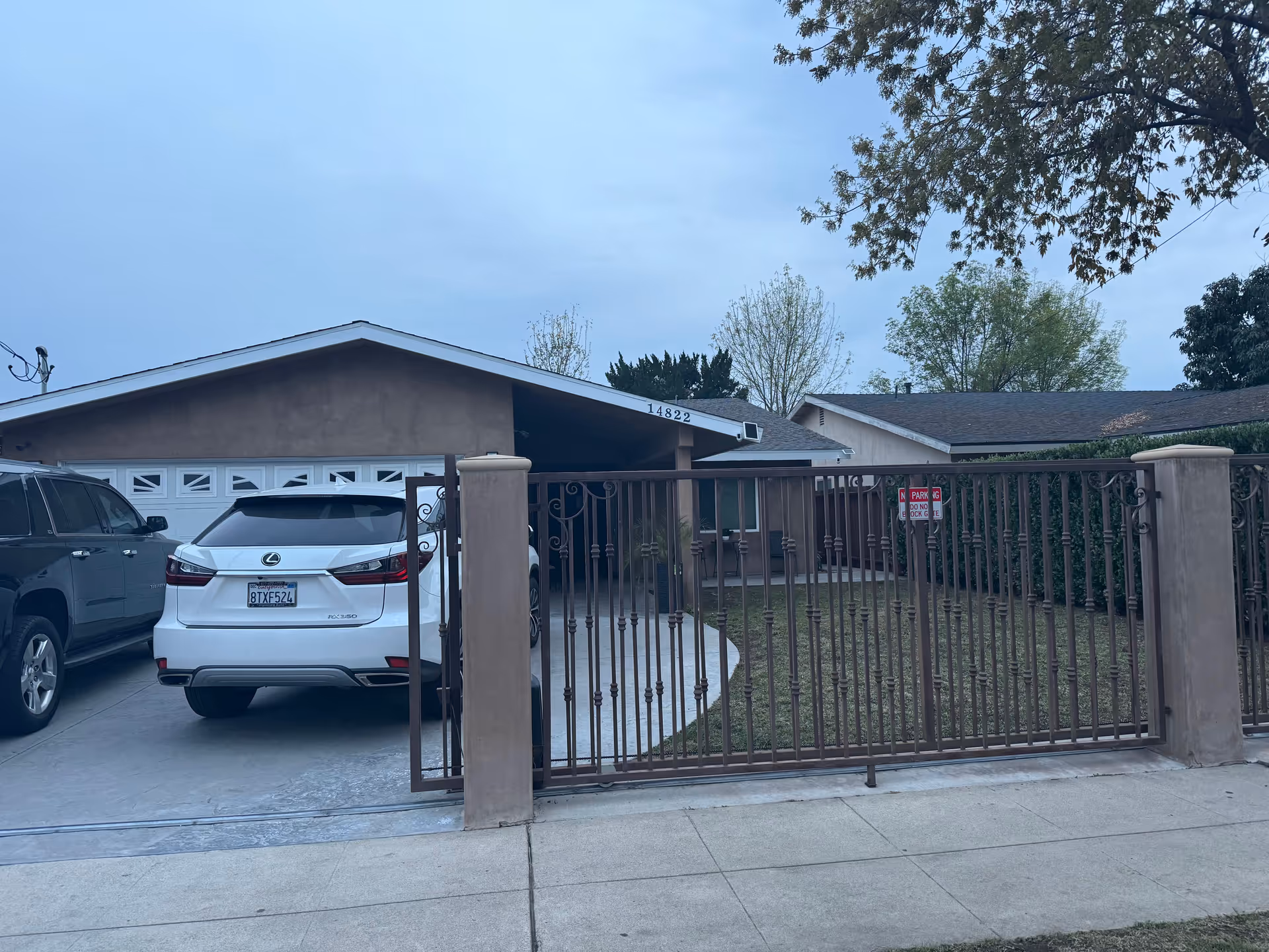 Front exterior view of a single-story residential building with a brown roof and beige walls, a driveway with two parked cars, and a metal gate enclosing a small front yard with grass and trees.