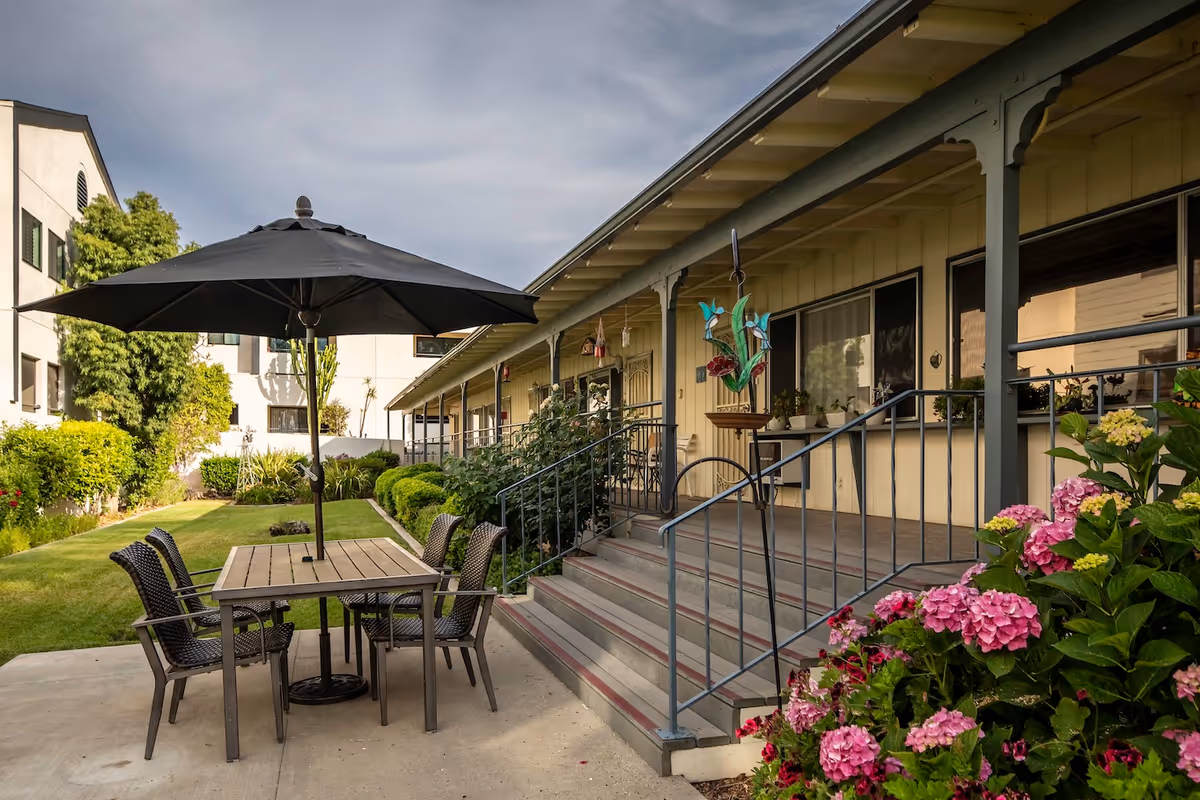 Outdoor patio area at Redwood Terrace featuring a table with four chairs and a large black umbrella. The patio is adjacent to a building with a covered porch and steps leading up to it. There are green bushes, pink flowers, and a well-maintained lawn surrounding the patio.