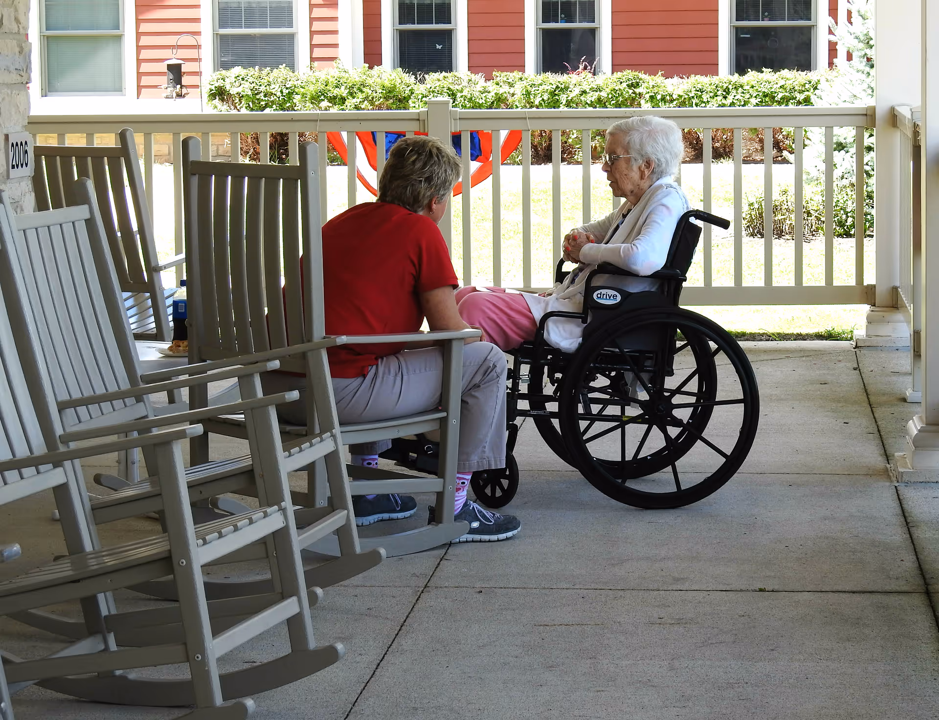 An elderly woman in a wheelchair talks with a caregiver seated on a covered porch beside several rocking chairs.