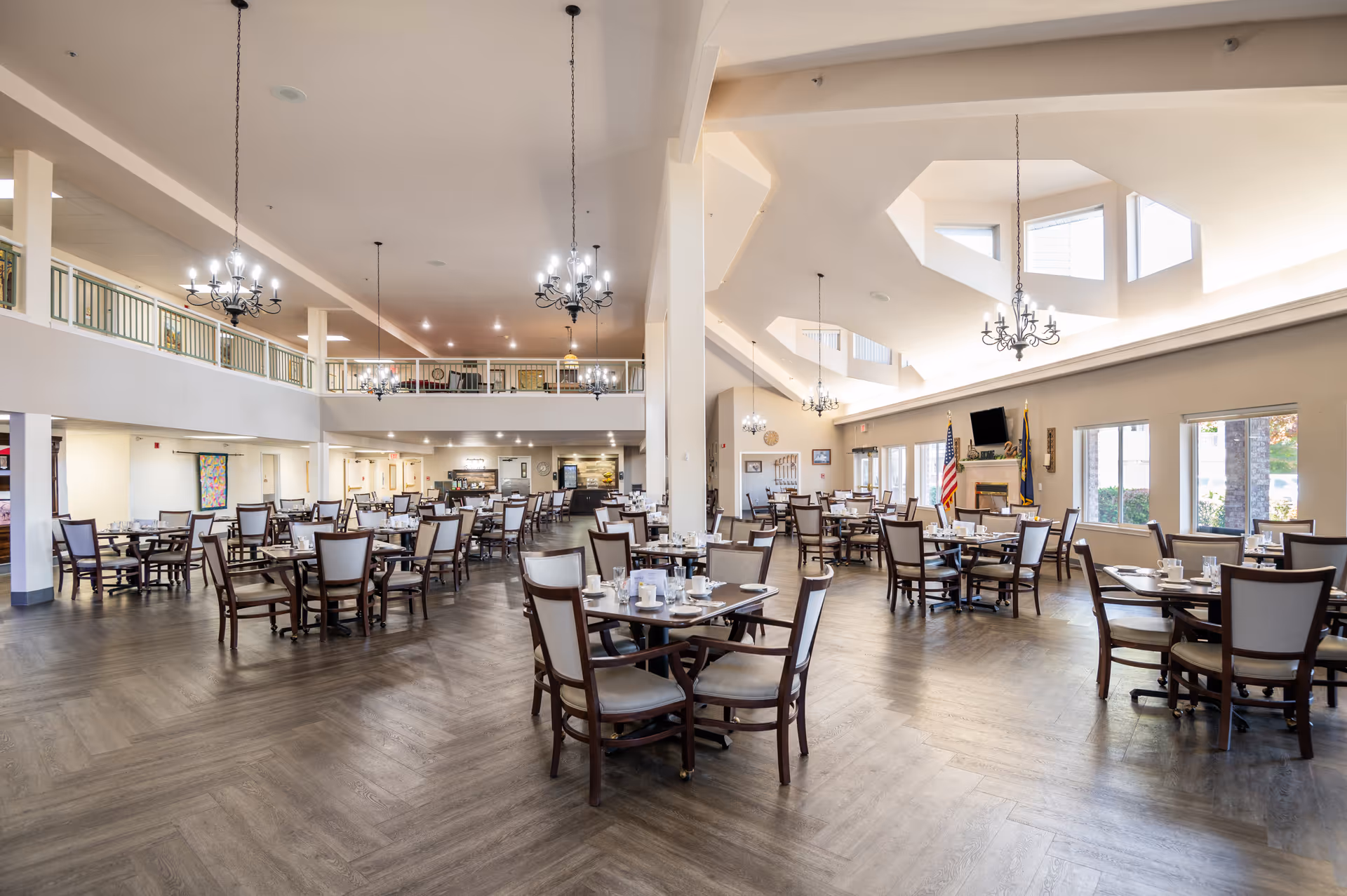 A spacious dining room in a senior living facility with multiple tables and chairs arranged neatly. The room features high ceilings with chandeliers, large windows allowing natural light, and a fireplace with a mounted TV above it. The floor has a wood pattern, and there is a balcony area overlooking the dining space.