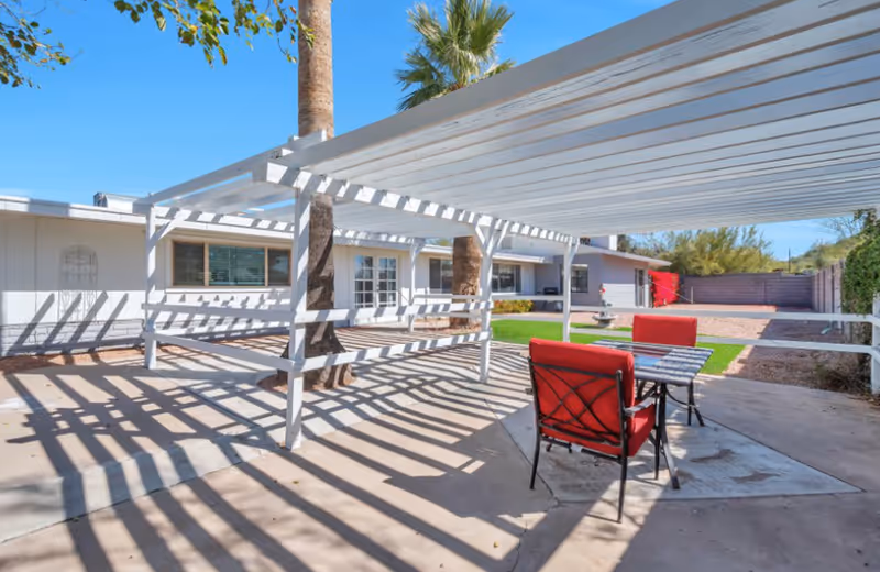 Outdoor patio area with a white pergola casting shadows on the concrete floor, two red cushioned chairs, and a small table. The background shows a single-story building with windows and a door, palm trees, green grass, and a clear blue sky.