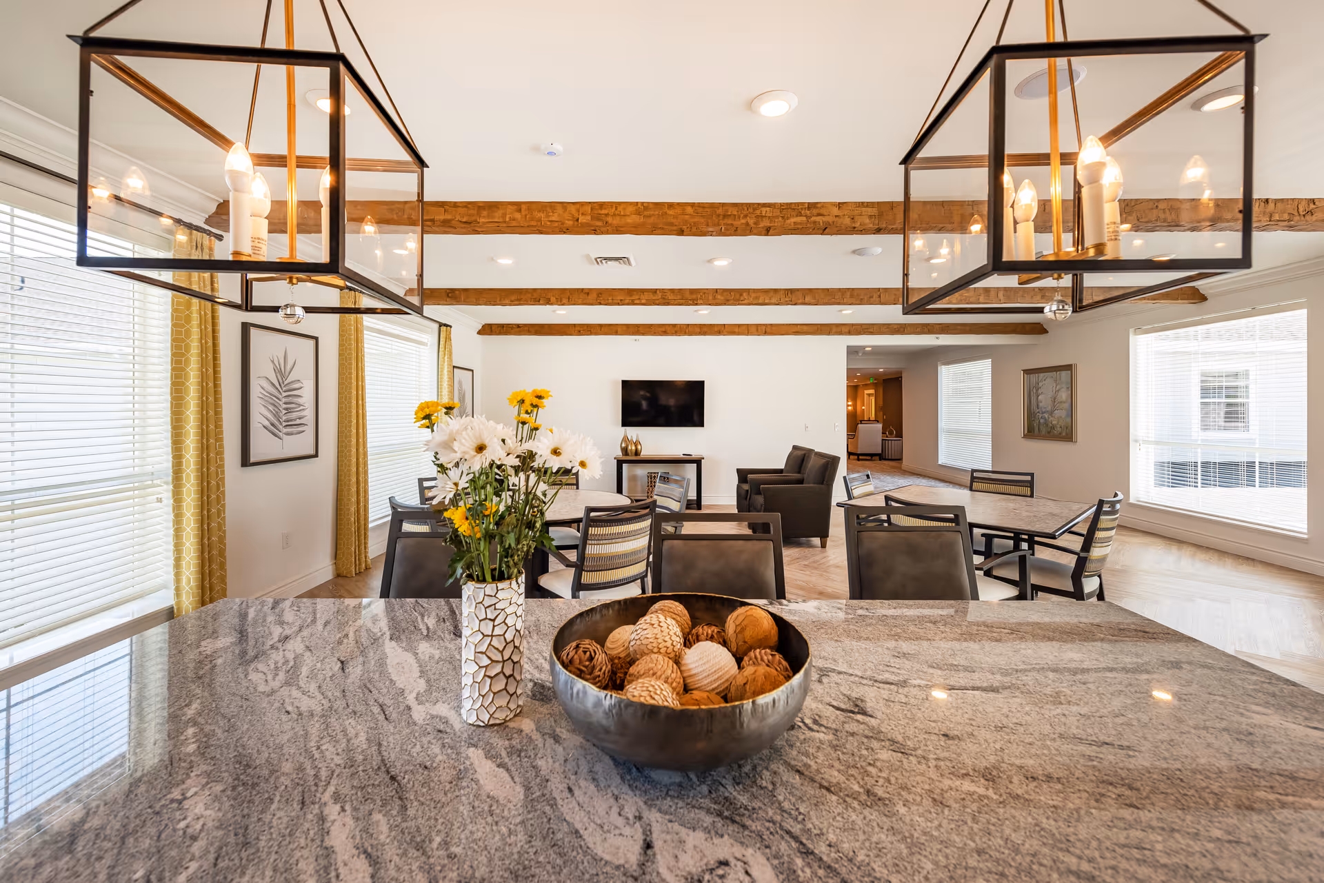 Bright and spacious common area with large windows and yellow curtains, featuring a granite countertop with a vase of white and yellow flowers and a decorative bowl with woven balls. The room has wooden beams on the ceiling, modern pendant lights, several tables and chairs, and a seating area with armchairs facing a wall-mounted TV.