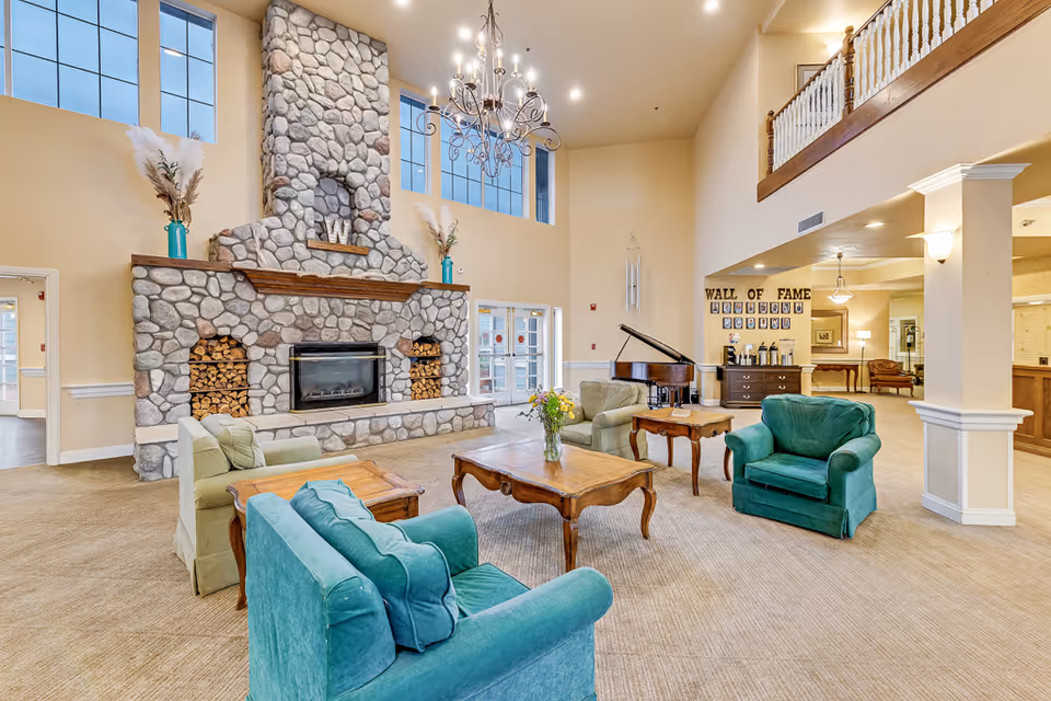 A spacious senior living facility common area with a large stone fireplace, teal and beige armchairs, wooden coffee tables, a grand piano, and a chandelier hanging from the high ceiling. There is a 'Wall of Fame' display on the far wall with framed photos.