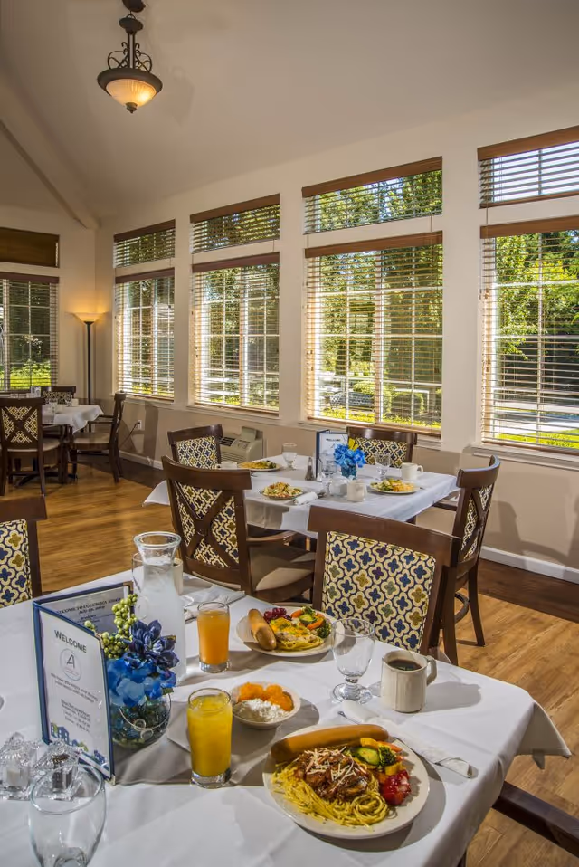 A bright dining room with large windows and wooden blinds letting in natural light. Several tables are set with white tablecloths, plates of food, glasses of juice, coffee cups, and decorative blue flowers. The room has wooden floors and patterned chairs around the tables.
