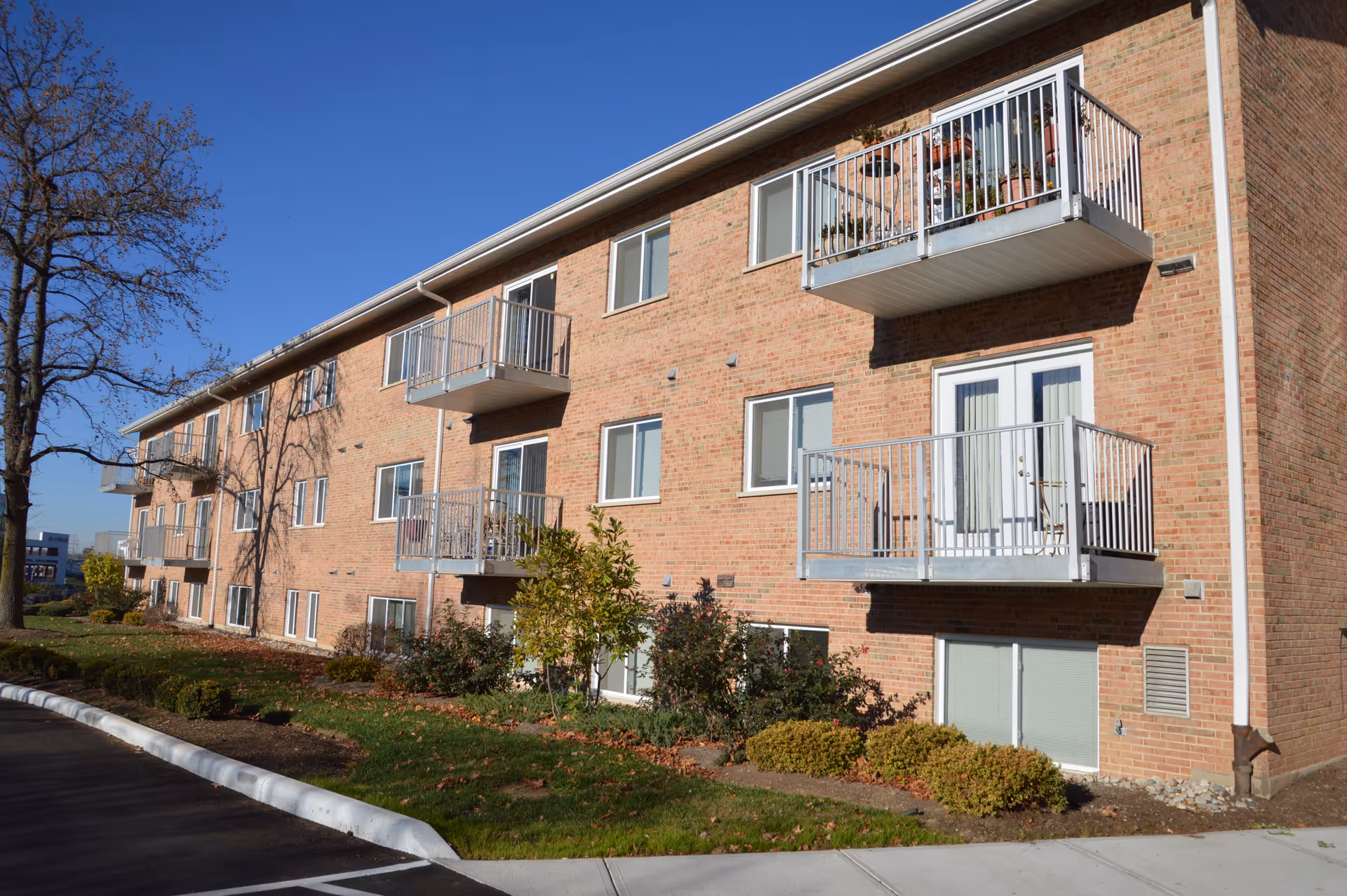 Exterior view of a three-story brick apartment building with multiple balconies and windows. There are small shrubs and trees planted along the building, and a clear blue sky overhead.
