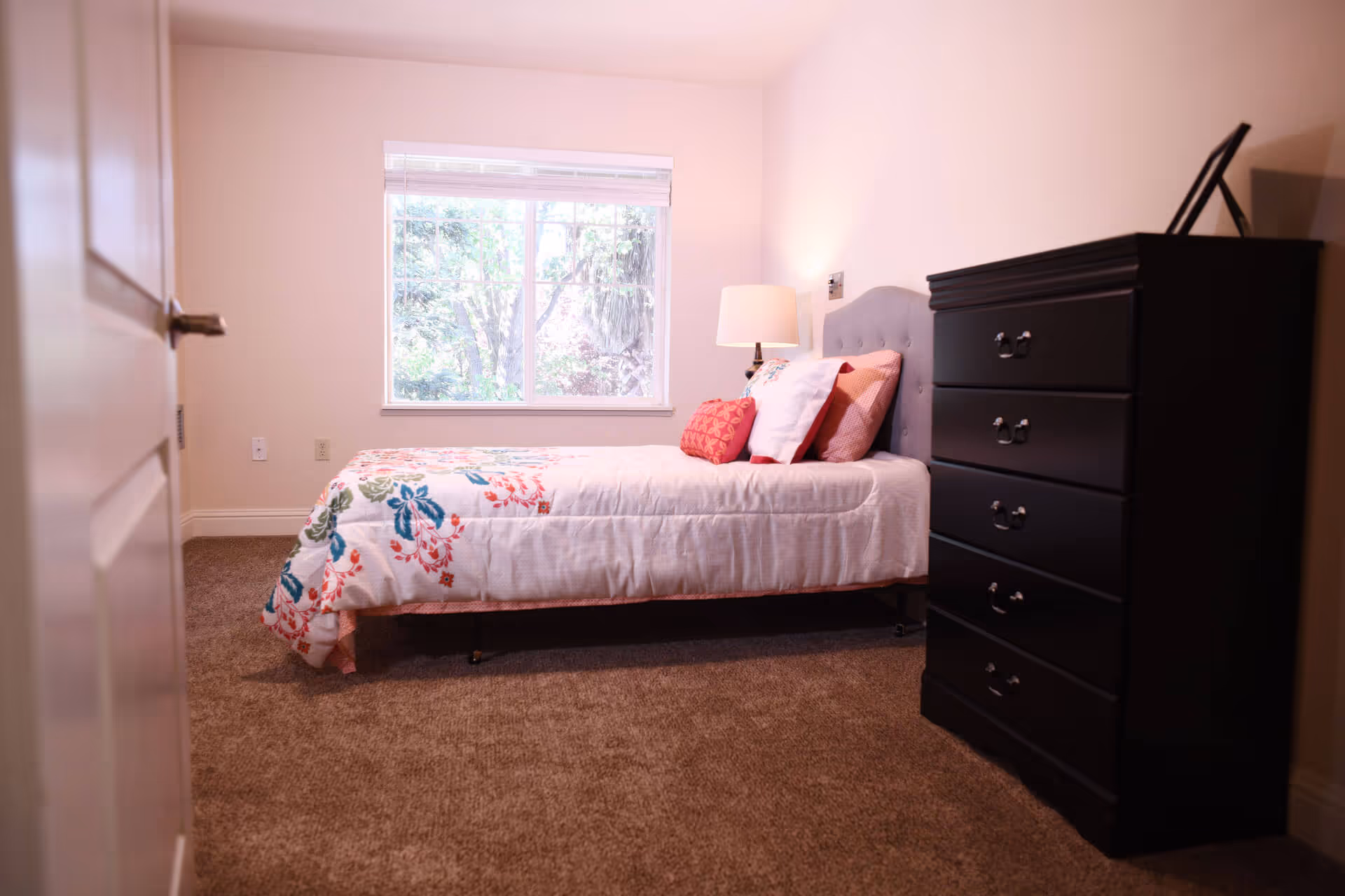 A cozy bedroom with a single bed featuring a floral patterned comforter and multiple pillows. Next to the bed is a bedside lamp on a small table. A large window with white blinds lets in natural light, and a dark wooden chest of drawers stands against the wall on the right. The room has beige carpet and light-colored walls.