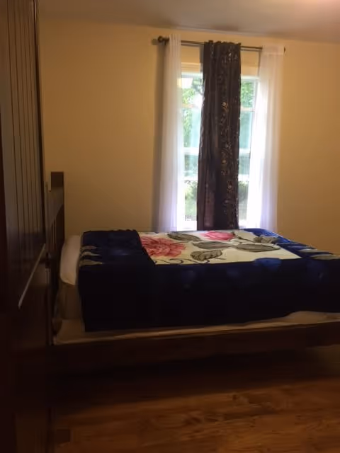 A small bedroom with a bed covered by a floral blanket and a dark blue throw. The bed is positioned against a wall with a window that has white sheer curtains and a dark decorative curtain in the center. The room has wooden flooring and light-colored walls.