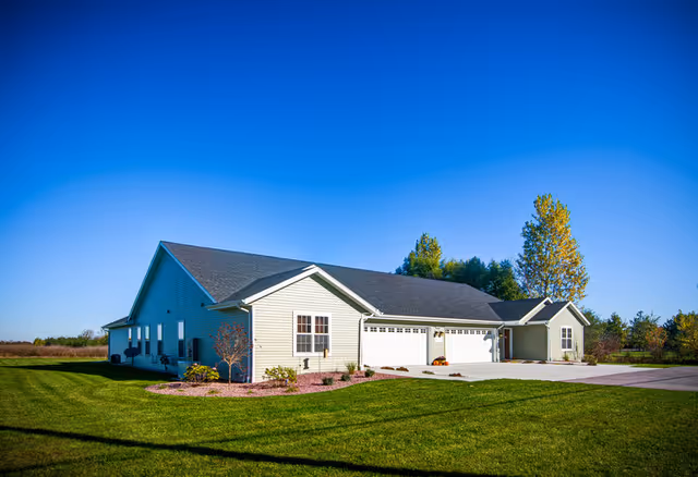 A single-story residential building with light-colored siding and a dark roof under a clear blue sky. The building has multiple garage doors and is surrounded by a well-maintained green lawn with some small trees and shrubs. Tall trees are visible in the background.
