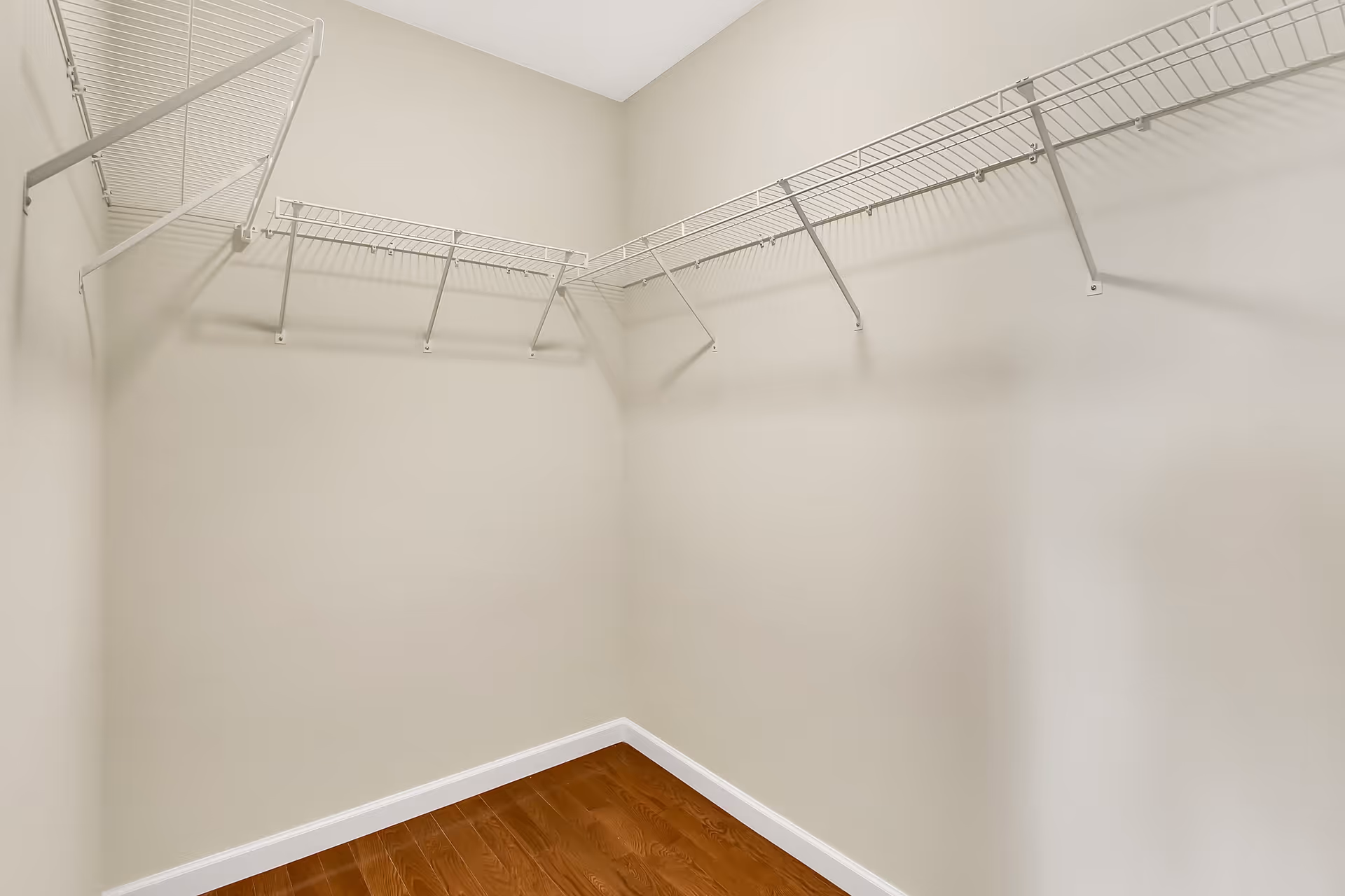 Empty walk-in closet with beige walls, wooden floor, and white wire shelving racks mounted on two walls.