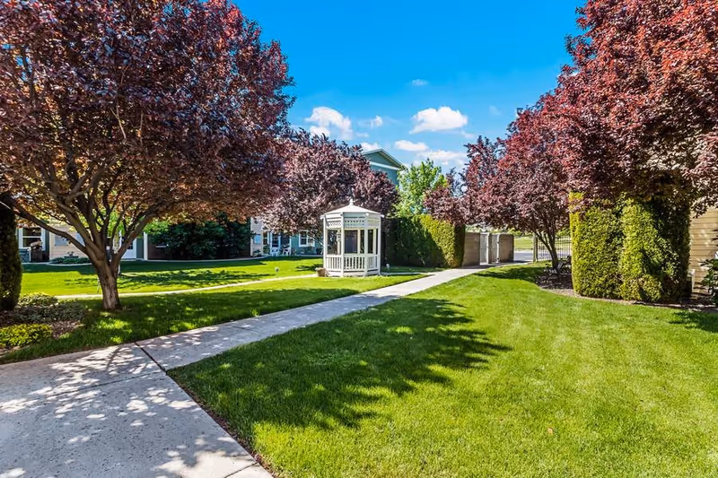 A well-maintained outdoor garden area with a concrete walkway leading to a white gazebo. The area is surrounded by lush green grass, neatly trimmed bushes, and trees with reddish-purple leaves under a bright blue sky with a few clouds.