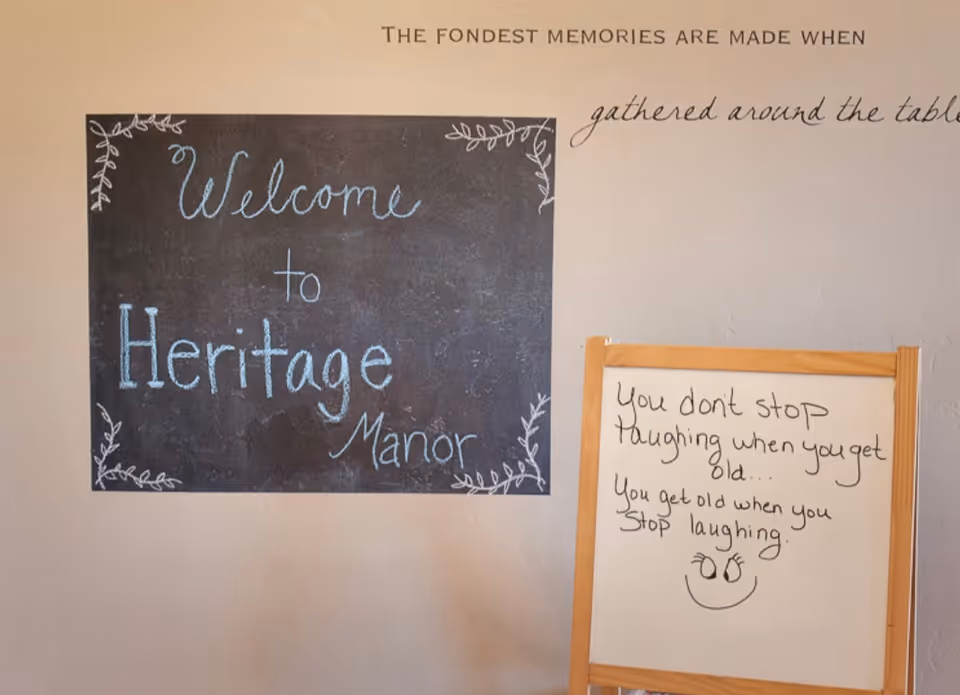A wall with a chalkboard sign that says 'Welcome to Heritage Manor' decorated with leaf designs, and a whiteboard with a handwritten quote: 'You don't stop laughing when you get old... You get old when you stop laughing.' Above the chalkboard, the wall has the phrase 'The fondest memories are made when gathered around the table.'