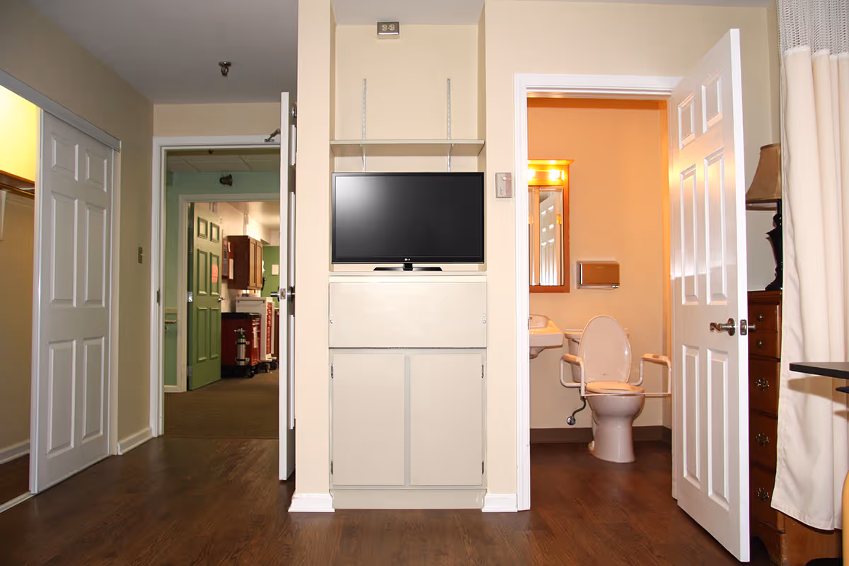 Interior view of a senior living room area with a wall-mounted TV cabinet centered and an open bathroom door revealing a toilet.