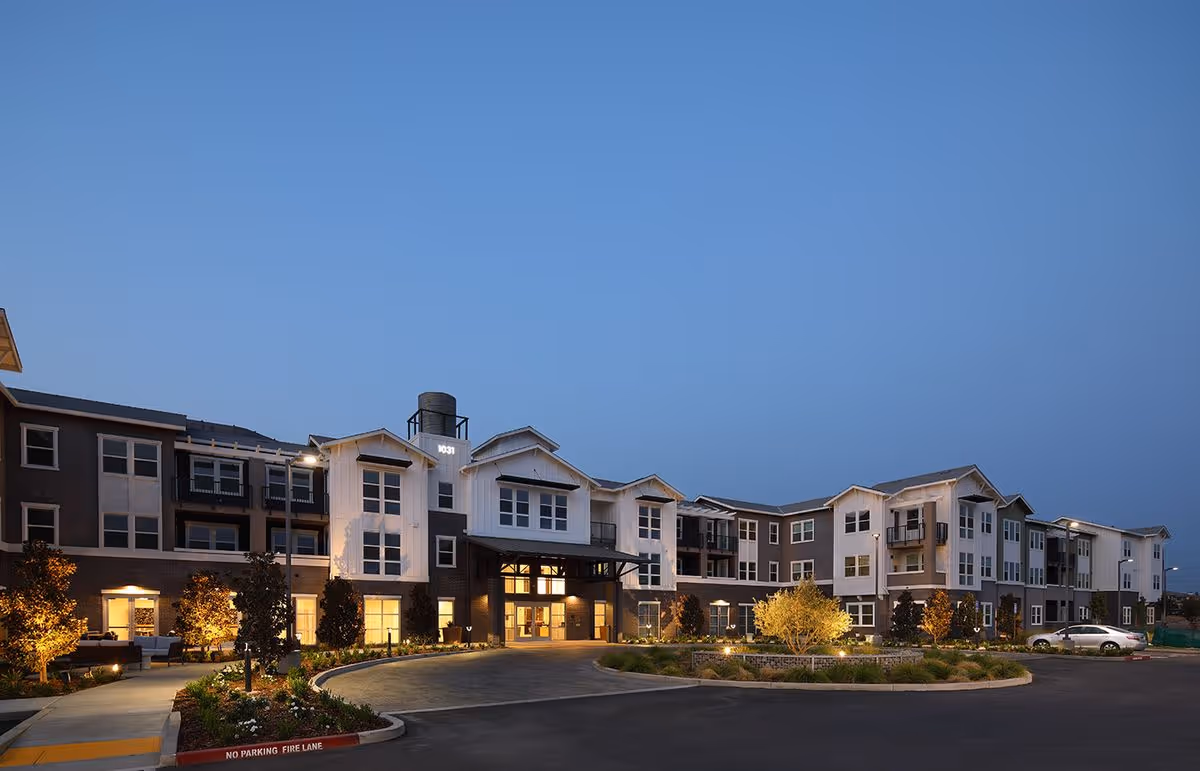 Front exterior of a three-story senior living building at dusk with a lit entrance, landscaped driveway, and parked cars.