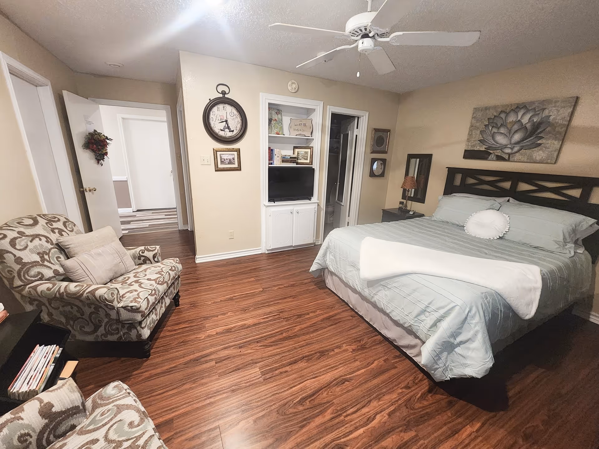 A cozy bedroom with a large bed covered in light blue bedding and a white round pillow. There is a wooden headboard and a floral painting above the bed. To the left, there is a patterned armchair with a pillow and a small bookshelf with magazines. The room has wooden flooring, a ceiling fan, and a built-in shelf with a TV and books. Two doors are visible, one leading to a hallway and the other to a bathroom.