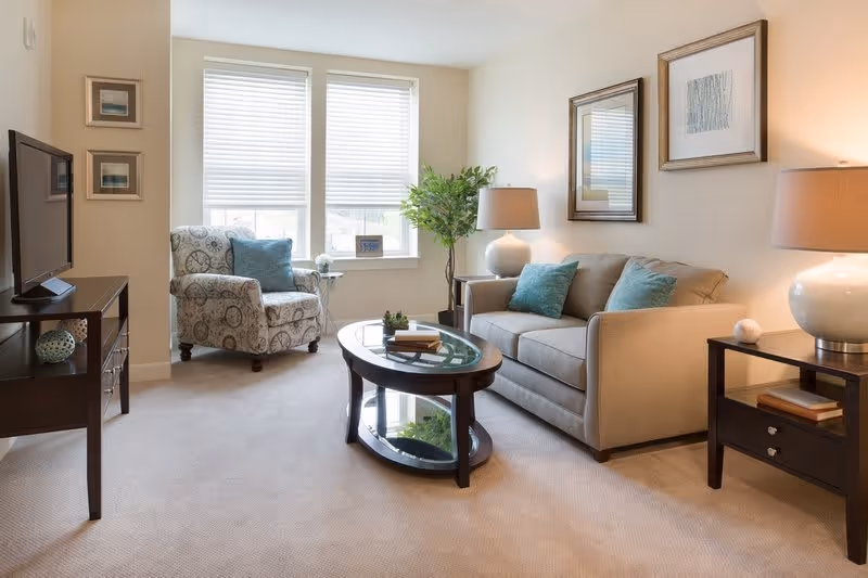 A bright and cozy living room with a beige sofa adorned with blue cushions, a patterned armchair, a glass-top coffee table, two side tables with lamps, framed artwork on the walls, and a TV on a stand. Two large windows with blinds allow natural light to fill the room.