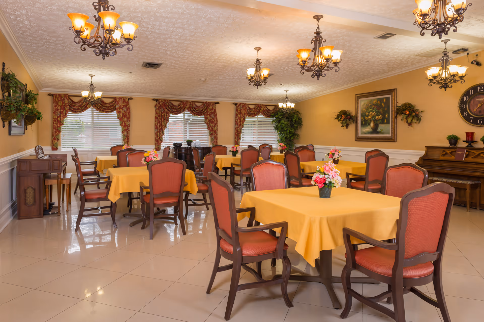 A well-lit dining room with multiple tables covered in yellow tablecloths, each adorned with a small flower arrangement. The room features red cushioned chairs, chandeliers hanging from the ceiling, large windows with floral curtains, a piano against the wall, and decorative plants and paintings.